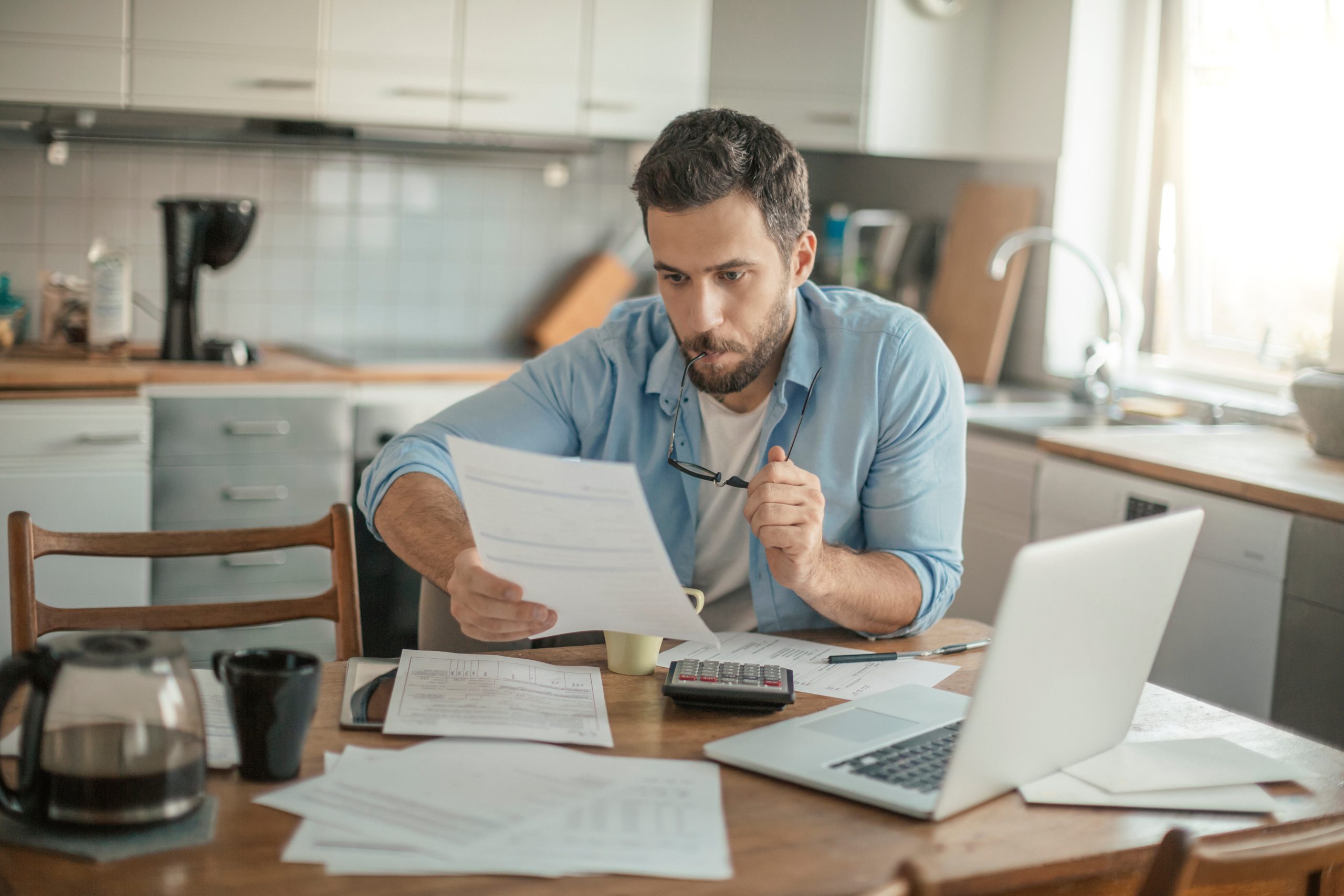 Comment financer sa formation en ligne avec INEAD. Jeune homme pensif analysant les financements possibles avec une calculatrice et des factures, dans un environnement familier, la cuisine d'une maison.